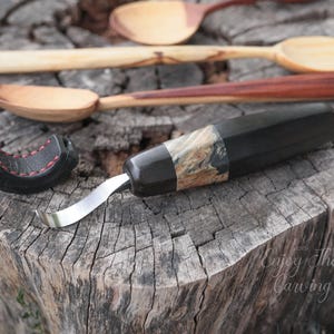 Könnte beinhalten: Nahaufnahme eines Holzschnitzwerkzeugs mit schwarzem und braunem Griff und silberner Klinge. Das Werkzeug liegt auf einer Holzoberfläche. Der Text "Enjoy the Carving" ist in der unteren rechten Ecke des Bildes sichtbar.