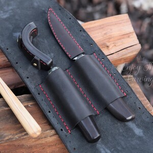 K&ouml;nnte beinhalten: Eine schwarze Lederrolle mit roten N&auml;hten, die f&uuml;r zwei Messer und ein Schnitzwerkzeug ausgelegt ist. Die Rolle hat eine Klappe, die mit einem Druckknopf geschlossen wird.