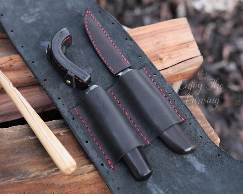 K&ouml;nnte beinhalten: Eine schwarze Lederrolle mit roten N&auml;hten, die f&uuml;r zwei Messer und ein Schnitzwerkzeug ausgelegt ist. Die Rolle hat eine Klappe, die mit einem Druckknopf geschlossen wird.
