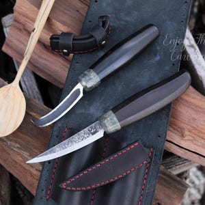 May include: Two handmade knives with wooden handles and black leather sheaths. The knives are on a black leather background with red stitching. The text "Enjoy The Carving" is visible in the background.