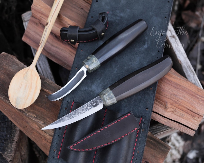 K&ouml;nnte beinhalten: Zwei handgefertigte Messer mit Holzgriffen und schwarzen Lederscheiden. Die Messer liegen auf einem schwarzen Lederhintergrund mit roten N&auml;hten. Der Text "Enjoy The Carving" ist im Hintergrund sichtbar.