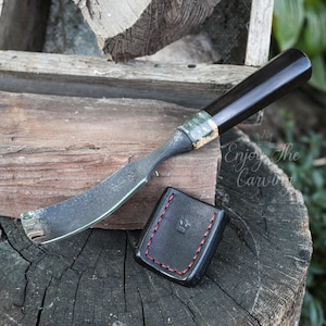 Puede incluir: Un cuchillo para tallar con una hoja curva y un mango negro descansa junto a una pequeña funda de cuero negro con costuras rojas. El cuchillo tiene una hoja plateada y un acento verde y dorado.