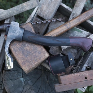 以下が含まれることがあります： 黒と赤ワイン色のハンドルが付いた手打ちの木彫り斧。斧は木製の表面に置かれ、赤いステッチの付いた革製の鞘が付いています。画像には「Enjoy the Carving」というテキストが表示されています。