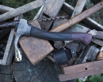 Può includere: Un'ascia per intagliare il legno forgiata a mano con un manico nero e bordeaux. L'ascia è appoggiata su una superficie di legno e ha un fodero in pelle con cuciture rosse. Il testo "Enjoy the Carving" è visibile nell'immagine.