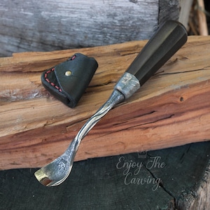 Puede incluir: Un primer plano de una herramienta de talla de madera con un mango de madera oscura y una hoja de metal. La herramienta está apoyada en un trozo de madera con una bolsa de cuero a su lado. El texto "Enjoy The Carving" es visible en la esquina inferior derecha de la imagen.