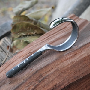 Puede incluir: Un gancho de metal con un mango curvo, hecho para colgar objetos. El gancho está pulido y tiene un acabado brillante. El gancho está apoyado en una pieza de madera.