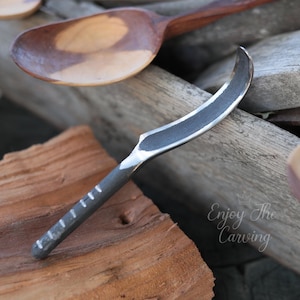 May include: A small, curved carving knife with a dark blade and handle, featuring a series of white markings. The knife is set against a backdrop of wooden planks and a wooden spoon. The text "Enjoy The Carving" is visible.