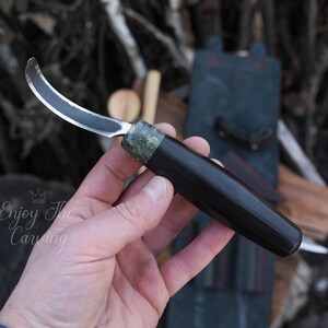K&ouml;nnte beinhalten: Ein gebogenes Schnitzmesser mit schwarzem Griff und einem gr&uuml;n-schwarzen, gemusterten Knauf. Das Messer wird in einer Hand gehalten.