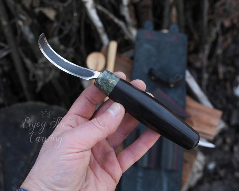 K&ouml;nnte beinhalten: Ein gebogenes Schnitzmesser mit schwarzem Griff und einem gr&uuml;n-schwarzen, gemusterten Knauf. Das Messer wird in einer Hand gehalten.