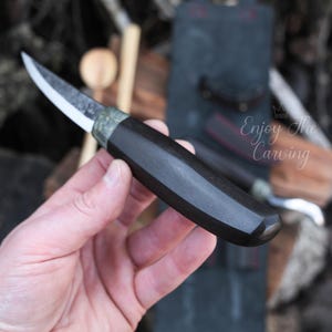 K&ouml;nnte beinhalten: Ein Schnitzmesser mit schwarzem Griff und einer gebogenen Klinge. Das Messer wird in einer Hand gehalten.