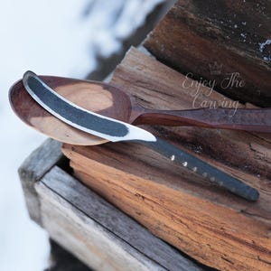 May include: A wooden spoon with a carved design rests on a piece of wood. A black and silver knife is placed on top of the spoon. The text "Enjoy The Carving" is visible on the wood.