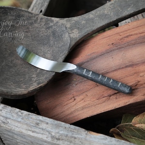 May include: A close-up of a small, silver, and black butter knife with a wooden handle. The knife is lying on a piece of wood with a wooden spoon in the background.