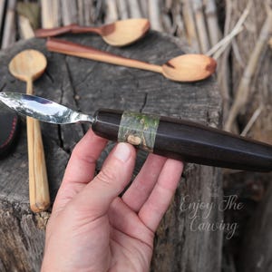 以下が含まれることがあります： 黒と緑のハンドルが付いた木彫りの道具のクローズアップ。道具は鋭く尖った刃が付いており、手で持たれています。背景には「Enjoy The Carving」というテキストが表示されています。