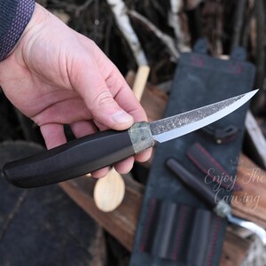 K&ouml;nnte beinhalten: Ein Messer mit schwarzem Griff, einer gebogenen Klinge und einer strukturierten Oberfl&auml;che. Das Messer wird in der Hand gehalten und die Klinge ist scharf. Das Messer ist Teil eines Sets von Schnitzwerkzeugen.