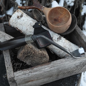 May include: A carving tool with a dark handle and a curved, silver blade rests in a wooden box with birch logs. A wooden cup and a spoon are also in the box. The text "Enjoy The Carving" is visible on the box.