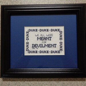 May include: A framed cross-stitch with a blue background and white text that reads "DUKE DUKE DUKE WE ALL WERE MEANT FOR DEVILMENT EBC 01 DUKE DUKE DUKE".