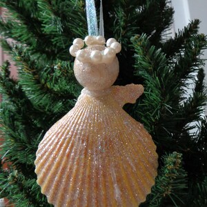 May include: A sparkly, beige seashell ornament with a pearl crown and a glittery finish. The ornament is hanging from a green artificial Christmas tree.