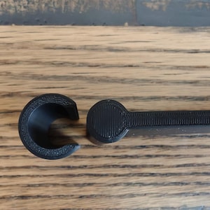 May include: A black, 3D-printed tool with a C-shaped end and a handle. The tool is made of a textured material and is designed for a specific purpose. The handle has a circular end. The tool is lying on a wooden surface.