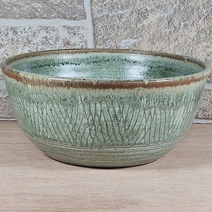 May include: A handmade ceramic bowl with a textured, carved design. The bowl features a green glaze with brown accents around the rim. The bowl is approximately 10 inches in diameter and 5 inches tall.