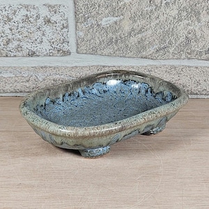 May include: A ceramic soap dish with a blue and green glaze. The dish is oval-shaped and rests on three small feet. The interior has a textured, mottled appearance.