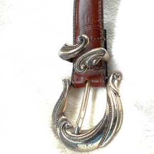 May include: Brown leather belt with a silver-colored buckle and belt loop. The buckle and loop have an ornate, swirling design. The belt is a medium brown color and appears to be made of leather.