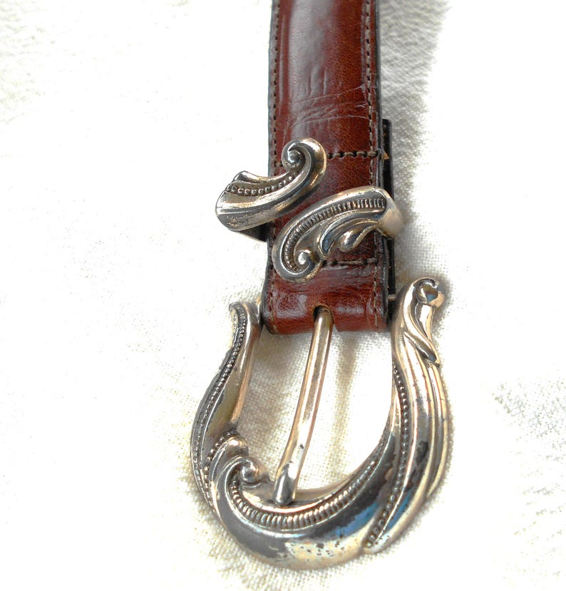 May include: Brown leather belt with a silver-colored buckle and belt loop. The buckle and loop have an ornate, swirling design. The belt is a medium brown color and appears to be made of leather.