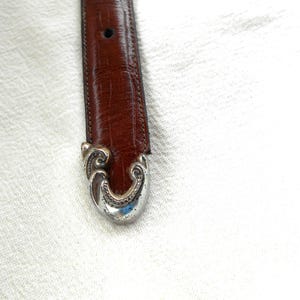 May include: Close-up of a brown leather belt with a decorative silver-colored buckle. The buckle has an ornate, swirling design. The belt is a medium brown color and appears to be made of genuine leather. The buckle is a bright silver color.