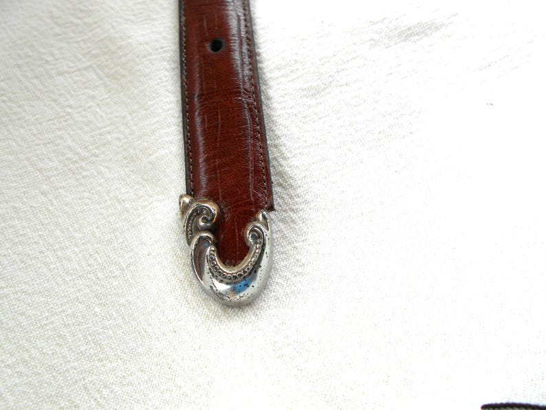 May include: Close-up of a brown leather belt with a decorative silver-colored buckle. The buckle has an ornate, swirling design. The belt is a medium brown color and appears to be made of genuine leather. The buckle is a bright silver color.
