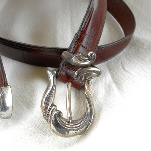 May include: A brown leather belt with a decorative silver buckle. The belt has a classic design with a detailed silver buckle and a matching silver end piece. The leather appears to be textured.