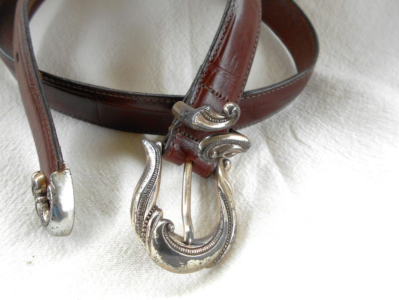 May include: A brown leather belt with a decorative silver buckle. The belt has a classic design with a detailed silver buckle and a matching silver end piece. The leather appears to be textured.
