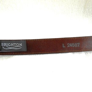 May include: A brown leather belt with a rectangular buckle. The belt has the brand name "BRIGHTON" printed on a black tag. The size is L 24507.