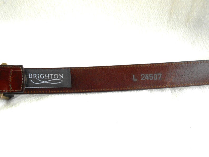 May include: A brown leather belt with a rectangular buckle. The belt has the brand name "BRIGHTON" printed on a black tag. The size is L 24507.