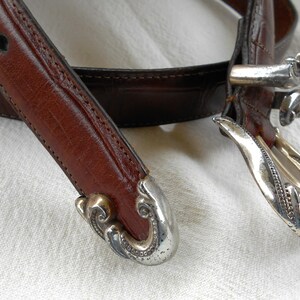 May include: Close-up of a brown leather belt with decorative silver-toned metal buckle and tips. The belt has a classic design with detailed stitching and a textured surface. The buckle and tips have an ornate, vintage style.