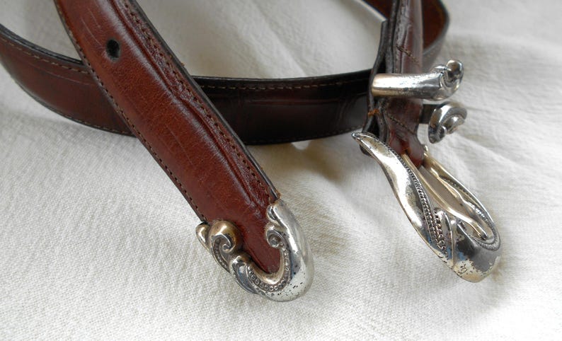 May include: Close-up of a brown leather belt with decorative silver-toned metal buckle and tips. The belt has a classic design with detailed stitching and a textured surface. The buckle and tips have an ornate, vintage style.