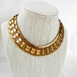 May include: A gold-tone metal choker necklace with a geometric design. The necklace is made of interlocking rectangular links.