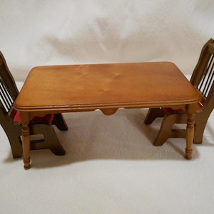 May include: A miniature wooden dining set featuring a rectangular table and two chairs. The table is light brown with a decorative apron. The chairs have a dark brown finish with red seat cushions. This set is ideal for dollhouses or miniature displays.