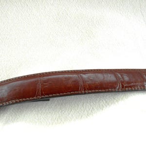 May include: A brown leather belt with a textured, crocodile-like pattern. The belt is a rich, dark brown color and appears to be made of high-quality leather. The stitching is visible along the edges.