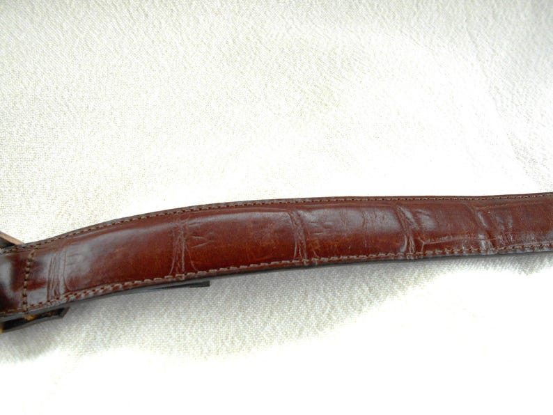 May include: A brown leather belt with a textured, crocodile-like pattern. The belt is a rich, dark brown color and appears to be made of high-quality leather. The stitching is visible along the edges.