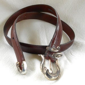 May include: A brown leather belt with a decorative silver buckle and matching silver accents. The belt is a rich, dark brown color and appears to be made of high-quality leather. The buckle has an ornate, swirling design.