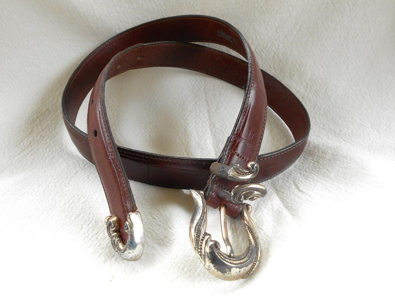 May include: A brown leather belt with a decorative silver buckle and matching silver accents. The belt is a rich, dark brown color and appears to be made of high-quality leather. The buckle has an ornate, swirling design.
