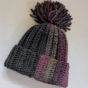 May include: A handmade crochet beanie in shades of gray, pink, and purple. The hat features a ribbed design on the gray section and a textured stitch on the pink and purple section. A large, multi-colored pom-pom tops the hat.