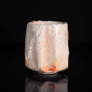 Könnte beinhalten: Handgefertigte, cremefarbene Keramiktasse mit strukturierter Oberfläche und einzigartiger, unregelmäßiger Form. Die Tasse hat dezente orangefarbene Akzente und steht vor schwarzem Hintergrund.