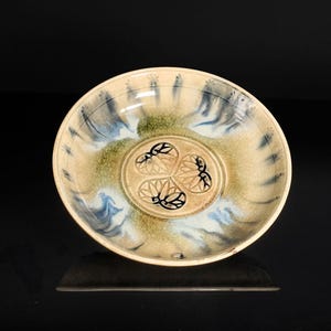 May include: A shallow ceramic bowl featuring a cream-colored base with blue and green glaze accents radiating from the center. The interior bottom displays a stamped design of three stylized circular floral motifs arranged in a triangular pattern.