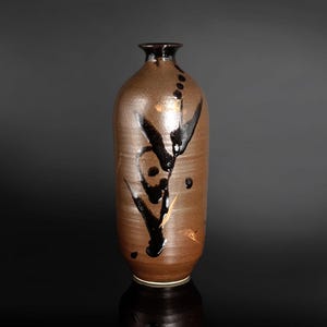 Może przedstawiać: Wysoki, cylindryczny wazon ceramiczny o matowym, brązowym wykończeniu. Powierzchnia ozdobiona jest wyrazistymi, abstrakcyjnymi czarnymi pociągnięciami pędzla, które owijają się wokół korpusu. Wazon ma wąską, rozszerzaną szyjkę i gładką, zaokrągloną podstawę.