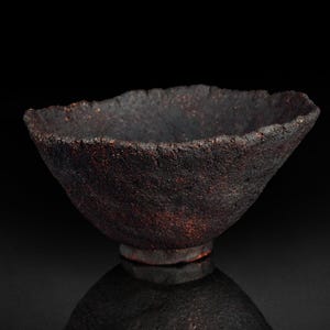 Puede incluir: Un cuenco de cerámica marrón oscuro hecho a mano con una superficie rugosa y texturizada. El cuenco tiene una forma irregular y orgánica con una amplia abertura y una pequeña base circular. El interior es oscuro y el exterior tiene acentos marrón rojizo.