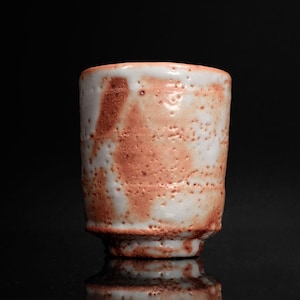 May include: A handmade ceramic cup with a unique textured surface. The cup features a white base with reddish-brown mottled patterns. The cup has a cylindrical shape with a slightly flared rim and a small footed base. The cup is approximately 3 inches tall.
