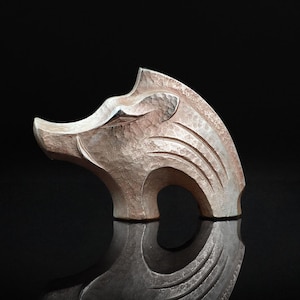 May include: A silver-colored sculpture of a boar's head with a textured surface and carved details. The artwork is set against a black background, with a reflective surface below the sculpture.