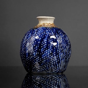 May include: A round ceramic vase with a blue and white crosshatch pattern. The vase has a small, off-white opening and a brown band at the neck. The vase is sitting on a reflective surface.