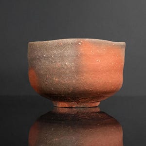 Puede incluir: Un cuenco de cerámica hecho a mano con una estética rústica. El cuenco presenta un diseño de dos tonos, con una mitad superior marrón oscuro y una mitad inferior rojo-anaranjado. La superficie del cuenco tiene un aspecto texturizado y se asienta sobre una pequeña base circular.