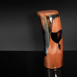 Puede incluir: Un jarrón alto de cerámica naranja con un esmalte negro y un diseño de corte único. El jarrón tiene un acabado liso y brillante y una forma ligeramente curvada.